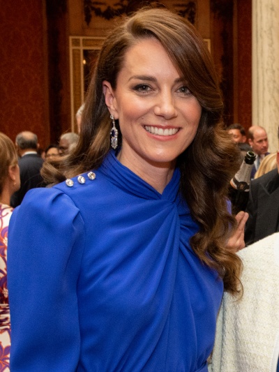 Prinzessin Kate in einer blauen Schluppenbluse. Ihre langen braunen Haare sind in Wellen gelegt und sie lächelt in die Kamera.