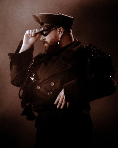 Sam Smith in einem Bühnenkostüm, das an eine Uniform erinnert. Er ist im Profil zu sehen, mit der Hand an der Militärmütze.