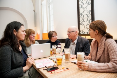 Vier Personen sitzen in einem Café um einen Tisch und sprechen miteinander. Auf dem Tisch steht ein aufgeklappter Laptop.