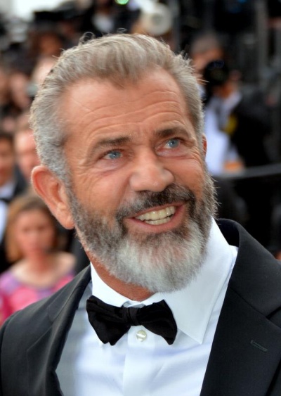 Mel Gibson mir grauen Haaren und Bart. Er trägt Anzug und Fliege und lächelt.
