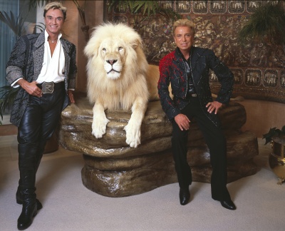 Siegfried und Roy stehen rechts und links neben einem weißen Tiger. Beide sind braun gebrannt und haben 80er-Jahre-Frisuren.