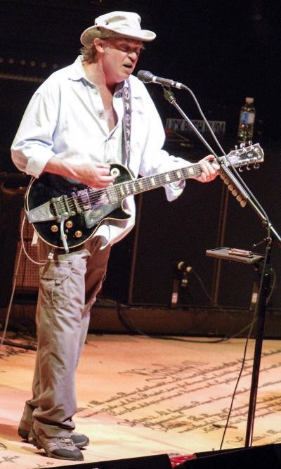 Neil Young singt auf der Bühne. Er spielt dabei Gitarre und trägt einen weißen Hut.