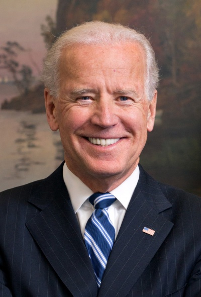 Joe Biden mit zurückgekämmten weißen Haaren. Er lächelt und trägt Anzug und Krawatte.