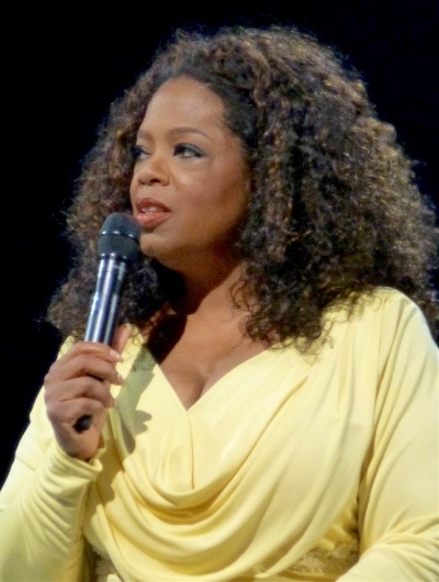 Oprah Winfrey mit dunkler Haut und schwarzen Locken. Sie trägt ein gelbes Langarmshirt mit Wasserfallausschnitt und spricht in ein Handmikro.