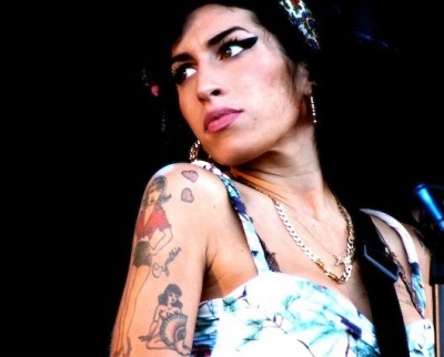 Amy Winehouse mit schwarzen, langen, zum Teil hochgesteckten Haaren mit starkem Make up in bunt gemusterter Kleidung