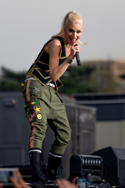 Gwen Stefani mit Hellblondem Pferdeschwanz in Hose und Weste auf einer Open Air Bühne. Sie singt lächelnd in ein Handmikro.