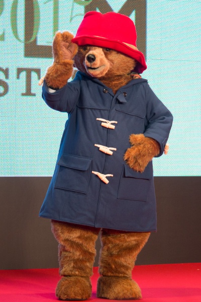 Ein Mensch in einem Kostüm als Paddington Bär. Er trägt einen roten Anglerhut und einen blauen Mantel. Er steht vor einer Logowand und winkt.