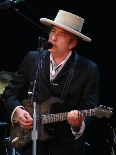 Bob Dylan spielt E-Gitarre und singt in ein Mikro. Er trägt Anzug und einen weißen Hut.
