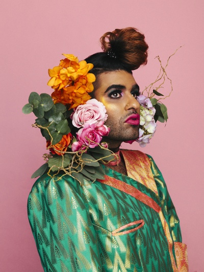 Alok in auffälliger bunter Kleidung und mit Blumen im Haar und am Kragen.