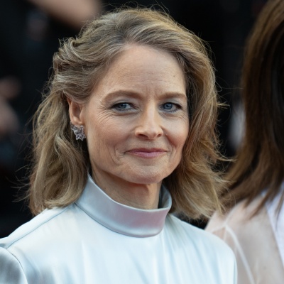Jodie Foster in einem eleganten weißen Kleid mit Turtle Neck. Ihre Haare sind gewellt und zum Teil hochgesteckt. Sie schaut mit einem leichten Lächeln in die Kamera.