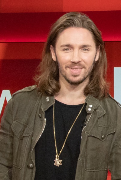 Gil Ofarim mit schulterlangen Haaren und einer Kette mit Davidstern. Er schaut direkt in die Kamera und lächelt leicht.