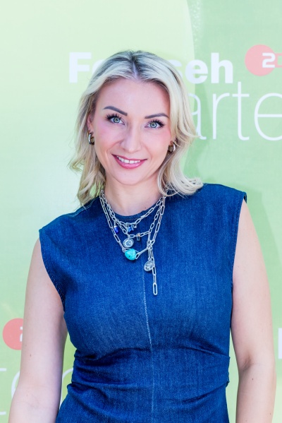 Anna-Carina Woitschack in einem Jeanskleid mit auffälligem Silberschmuck vor der Logowand des ZDF Fernsehgartens