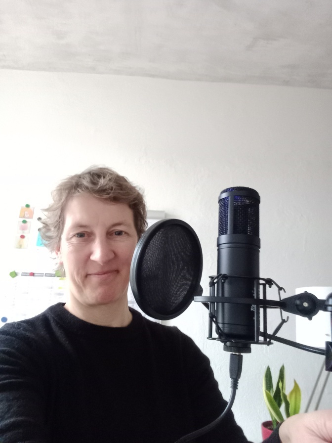 Eine Frau mit kurzen blonden Haaren sitzt vor einem Mikrofon und lächelt in die Kamera.